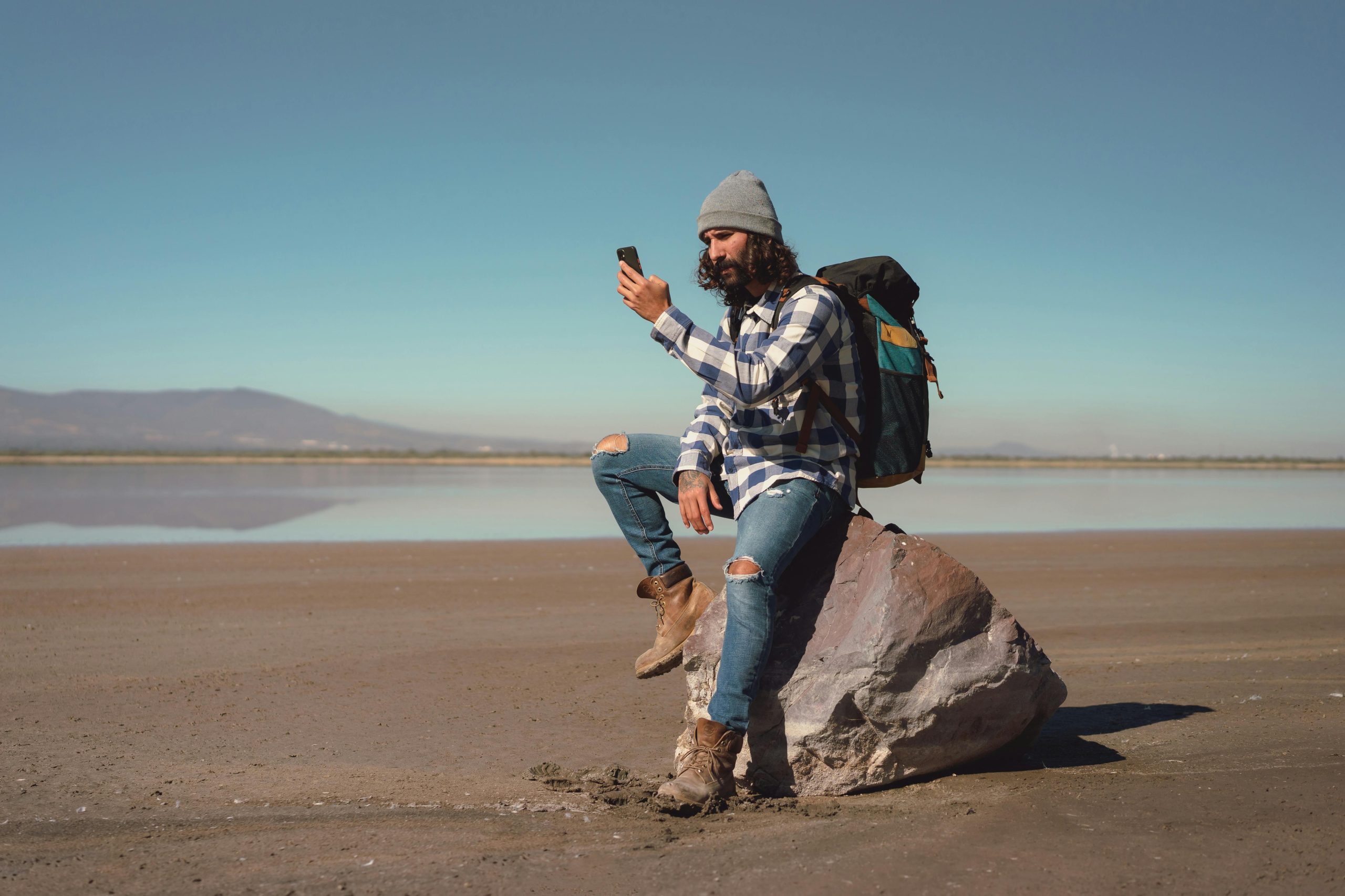 Man sitting on a rock by a serene lake, checking his smartphone while hiking in style.