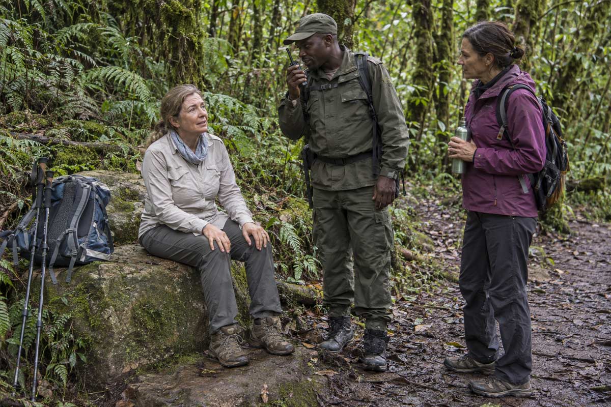 Guide using a handheld radio beside a seated trekker and her companion on a forest trail.